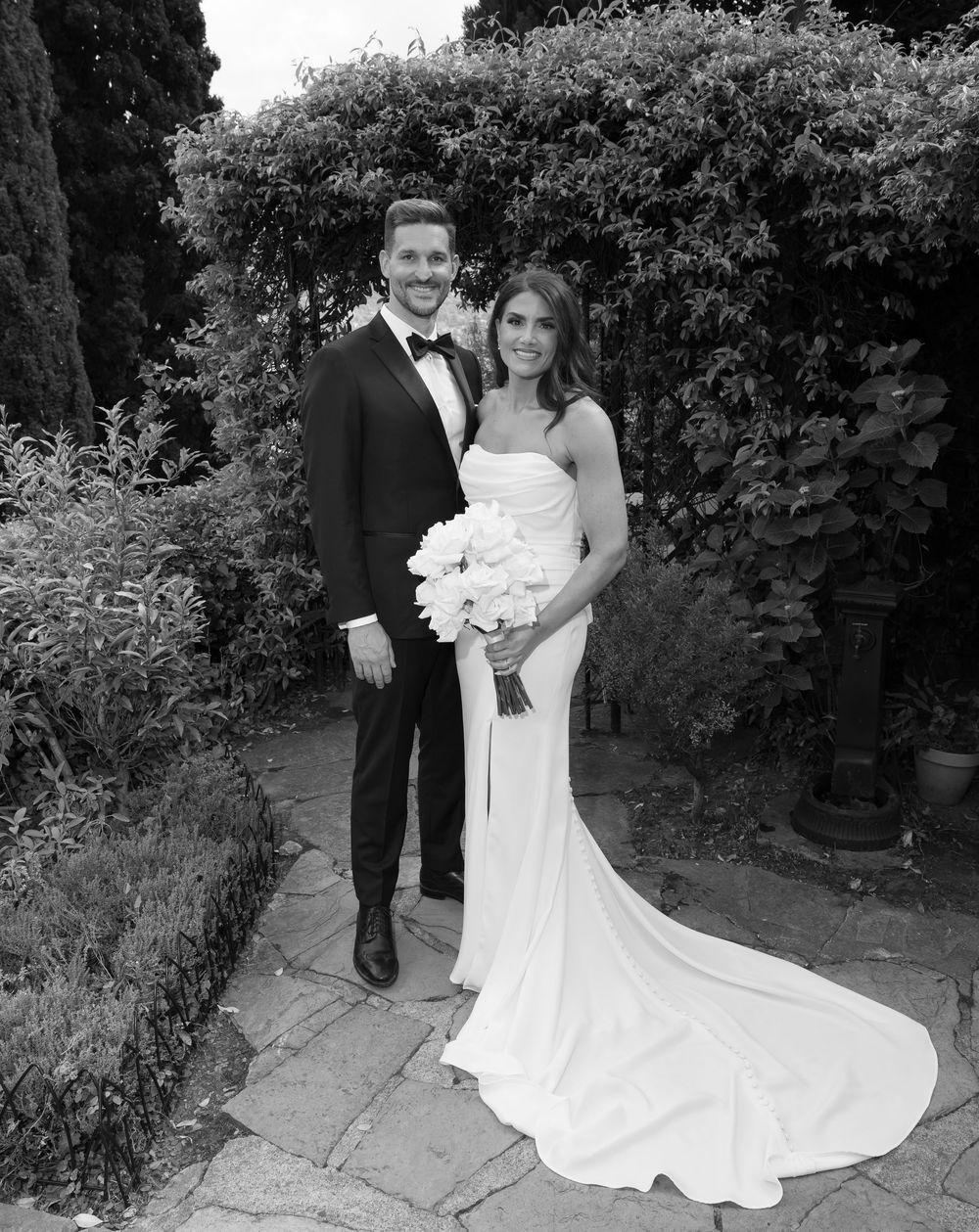 A wedding couple poses together in a garden pathway against lush greenery in a black and white photograph.