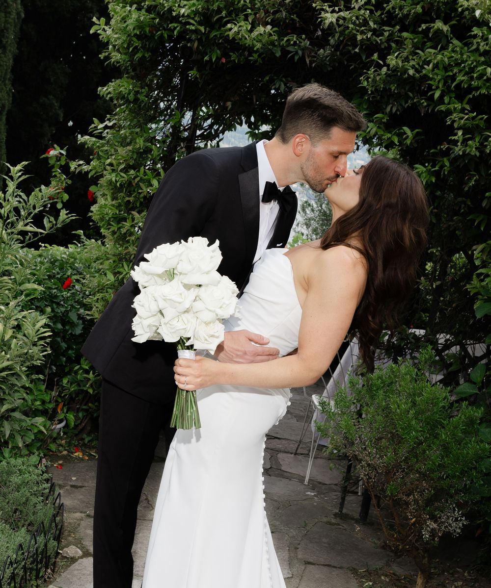 A romantic wedding couple shares a kiss in a garden setting while the bride holds a bouquet of white flowers.