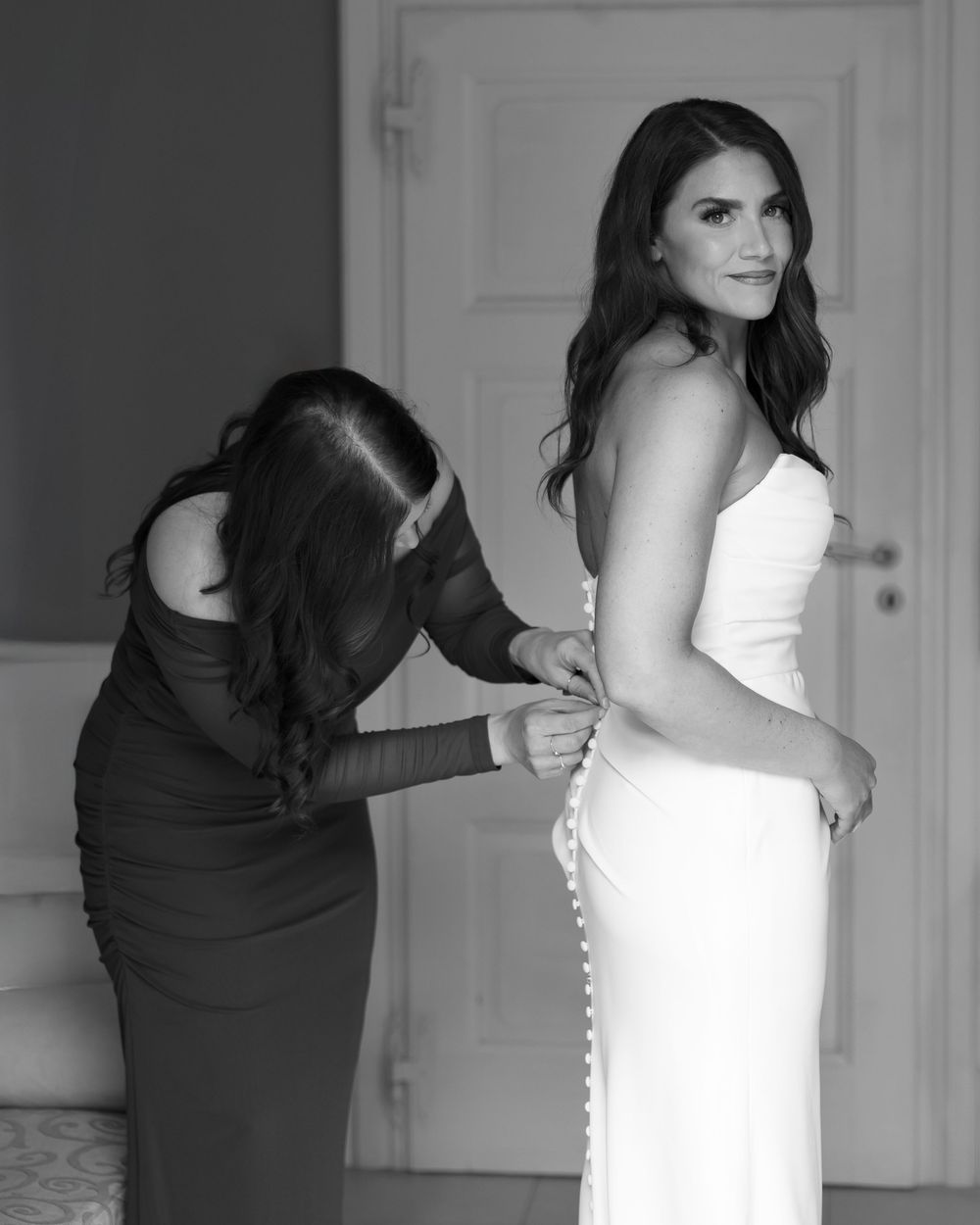 A woman in a white wedding dress stands while someone helps adjust and zip up the back of her gown.