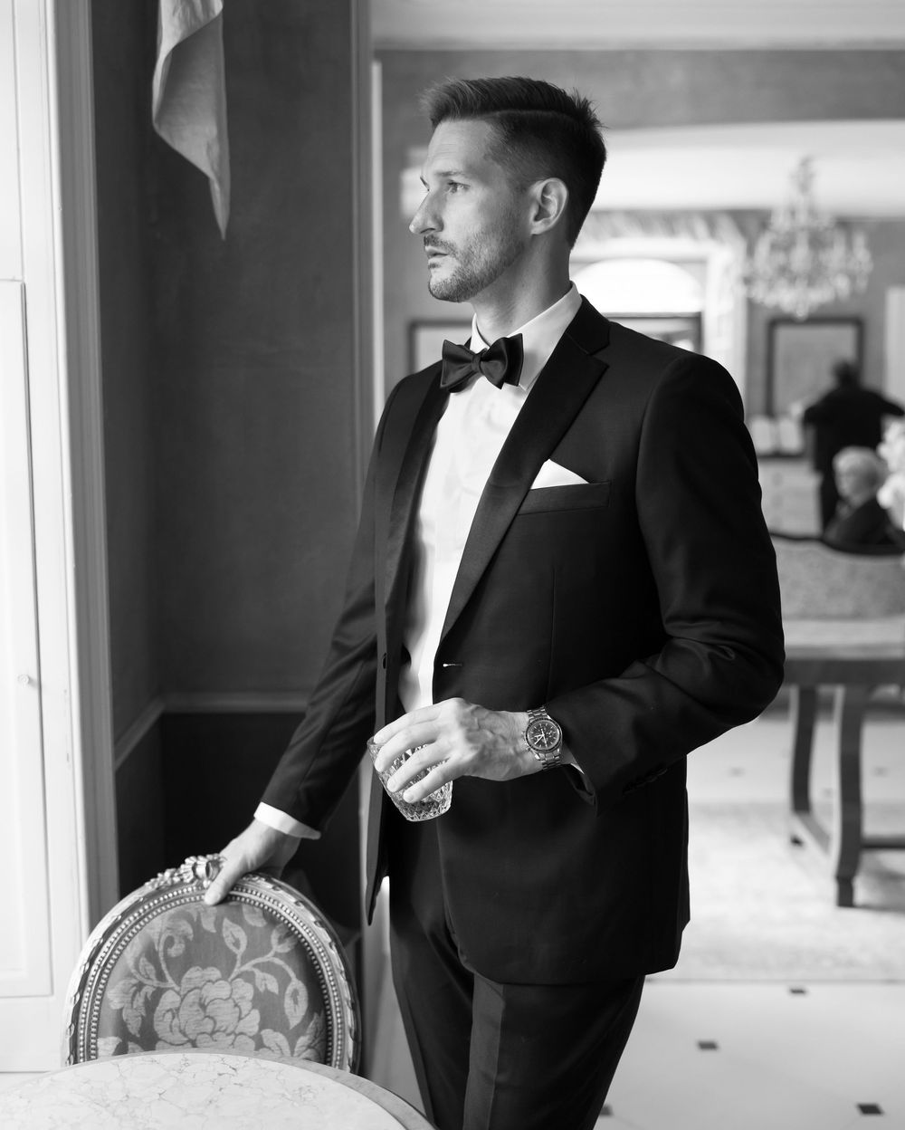 A person in a classic black tuxedo and bow tie stands elegantly beside an ornate chair in a formal black and white portrait.