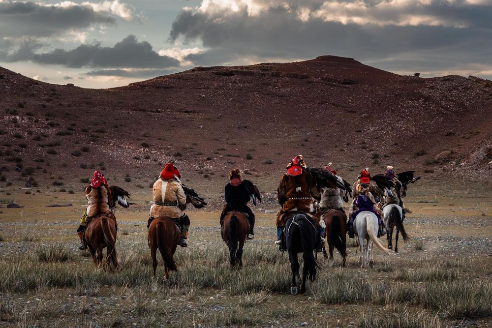 A group of horse riders traverse across a rugged mountainous landscape at sunset.
