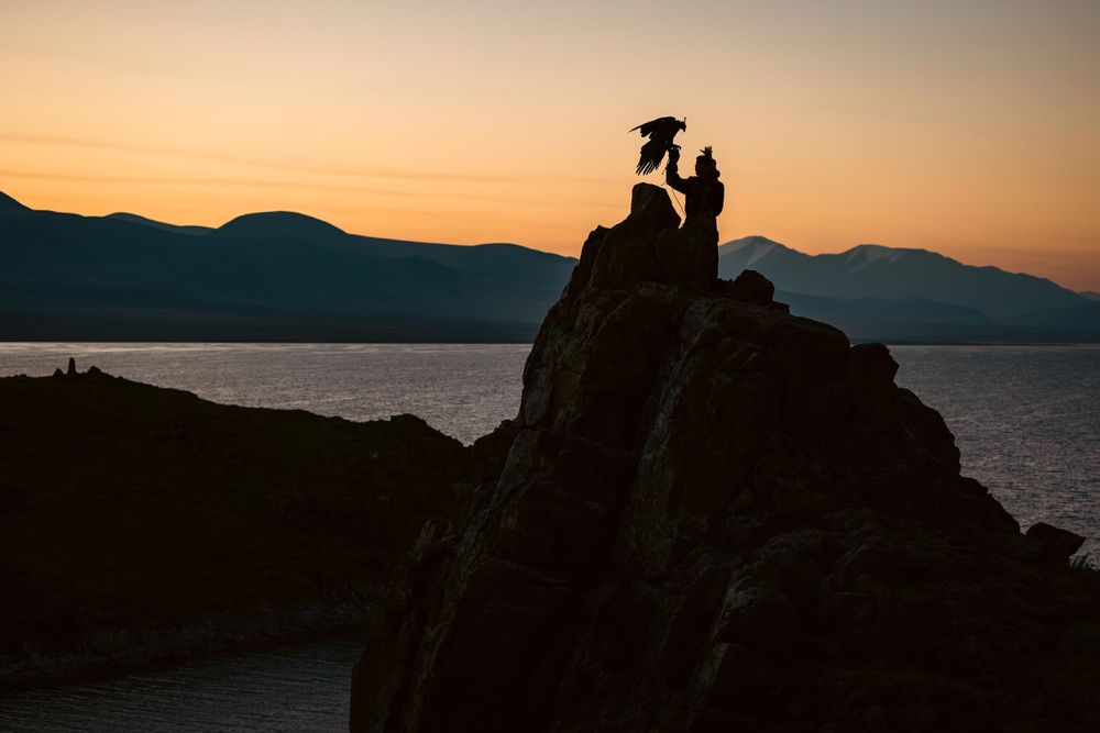 A silhouette of two figures on a rocky cliff overlooking a lake at sunset.