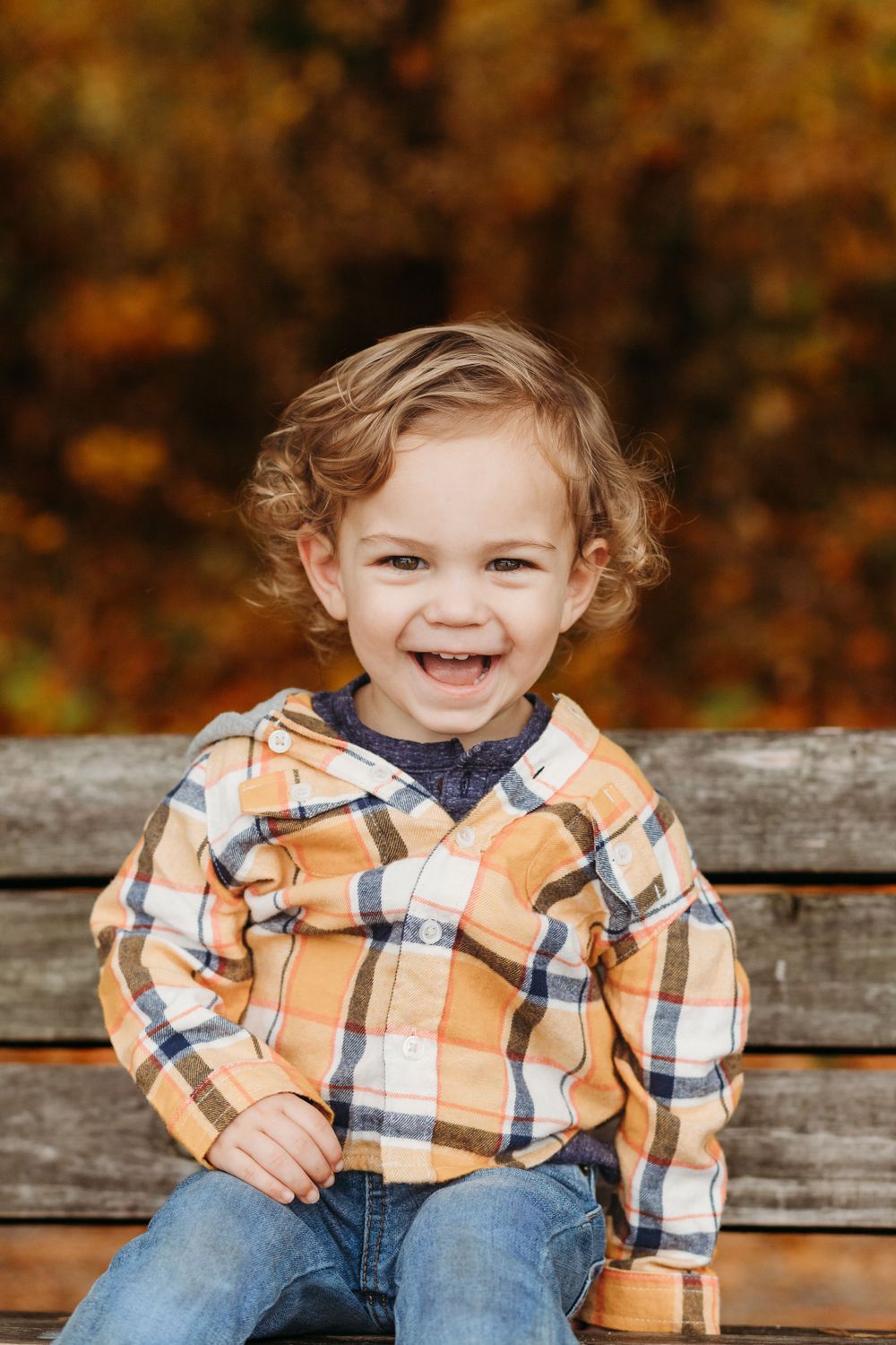 Child in plaid orange flannel shirt and jeans sits on wooden bench with autumn foliage in background.