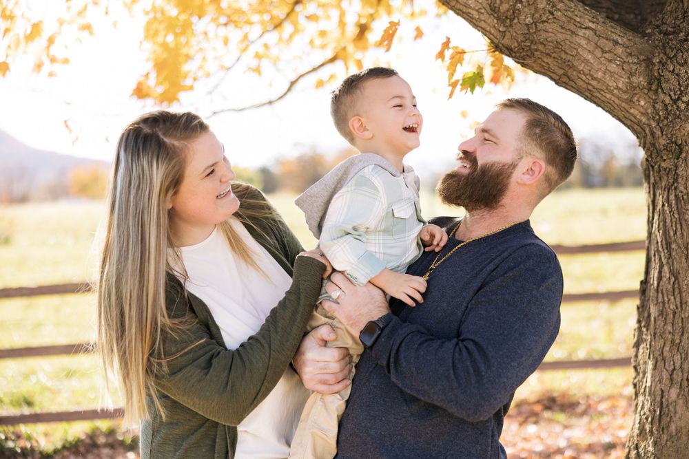 A family shares laughter and joy together under an autumn tree with a rustic fence in the background.