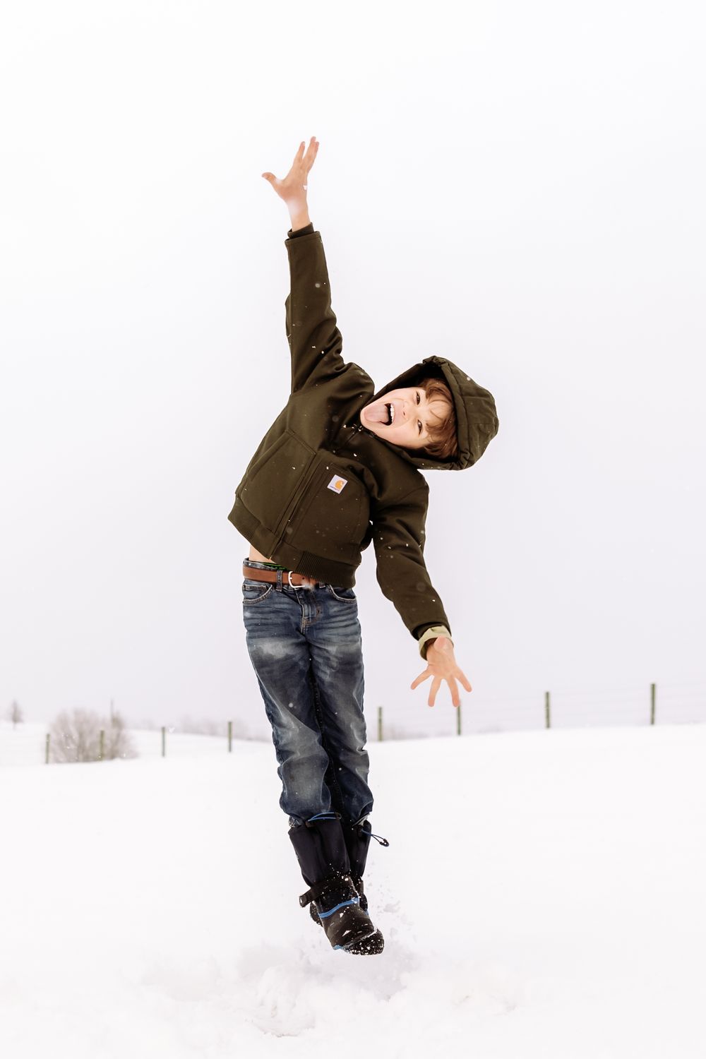A person in a brown jacket and jeans jumps playfully in the snow against a white winter background.
