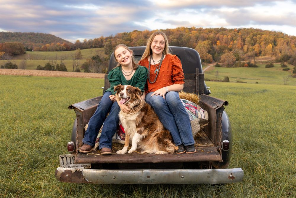 Two young people sit with an Australian Shepherd mix dog in the bed of a pickup truck in a scenic autumn field.