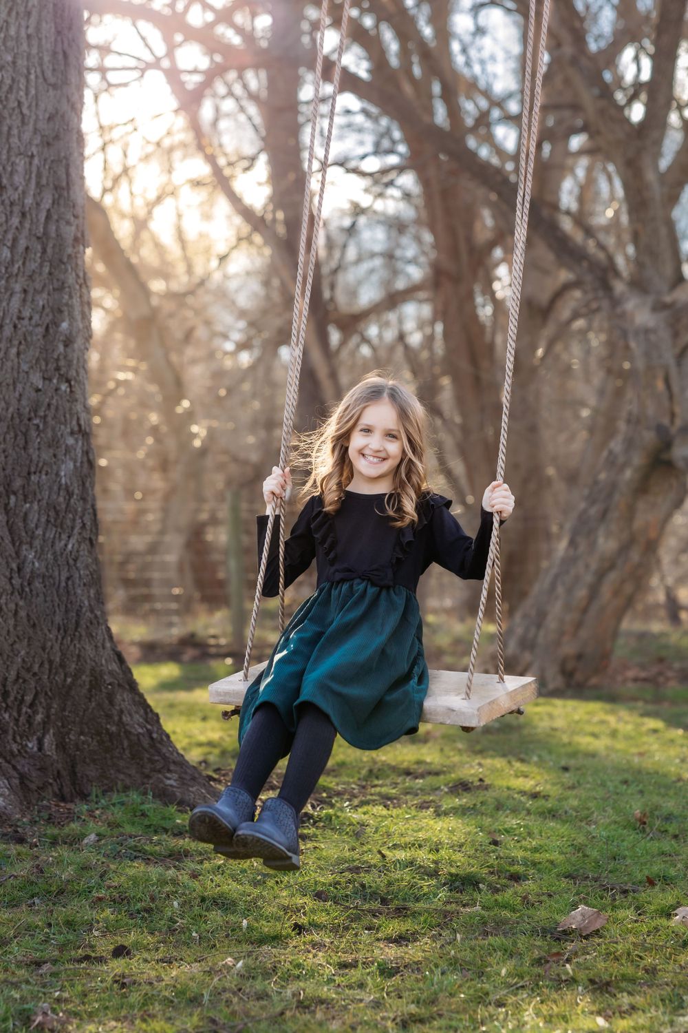 A young child in a dress and boots enjoys swinging on a tree swing at sunset in a wooded park.