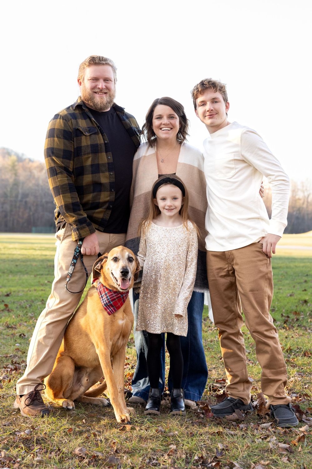 A family poses together outdoors in a grassy field with their tan dog wearing a red bandana.