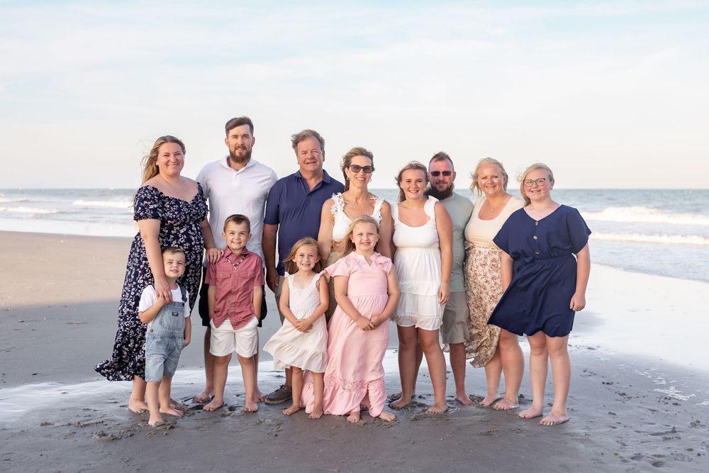 Multiple families pose on the beach in coordinated pastel outfits during sunset for a group photo session.