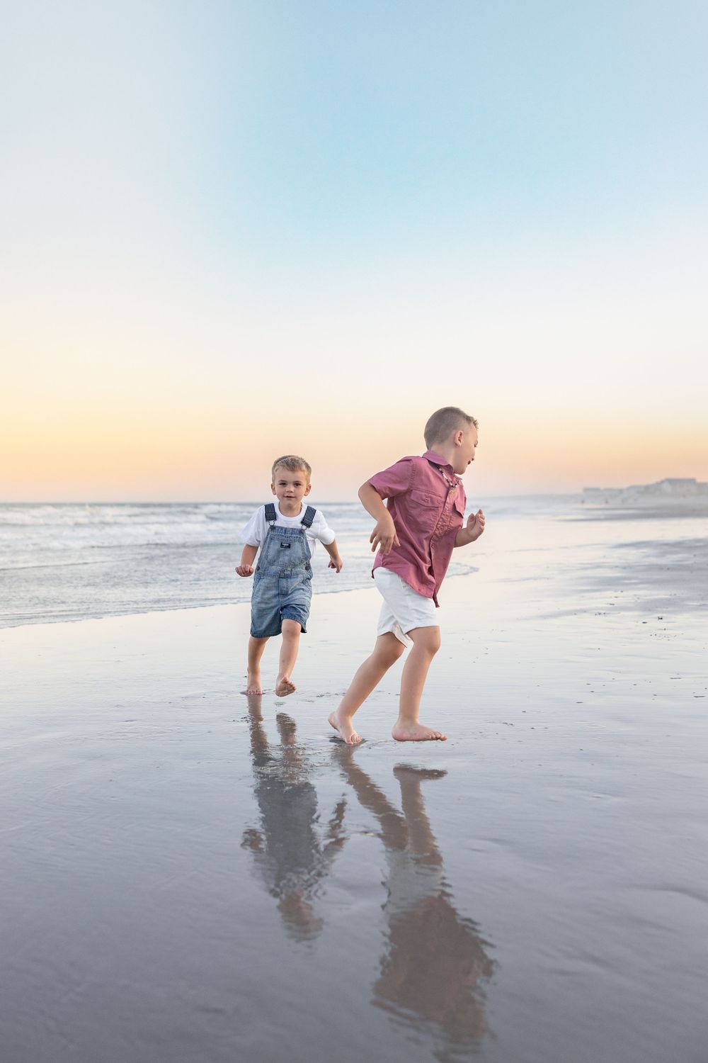 Two children run and play along a wet sandy beach at sunset with their reflections visible in the sand.