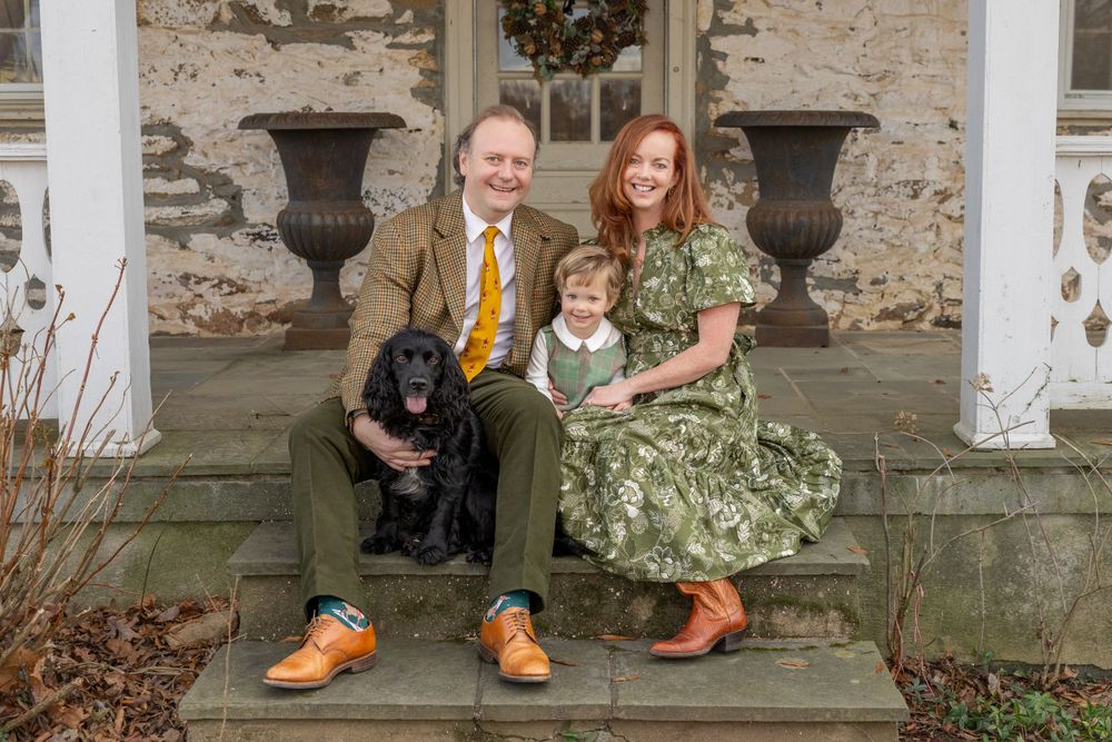 A couple in vintage-inspired clothing sits on stone steps with their black dog, against a textured stone wall backdrop.