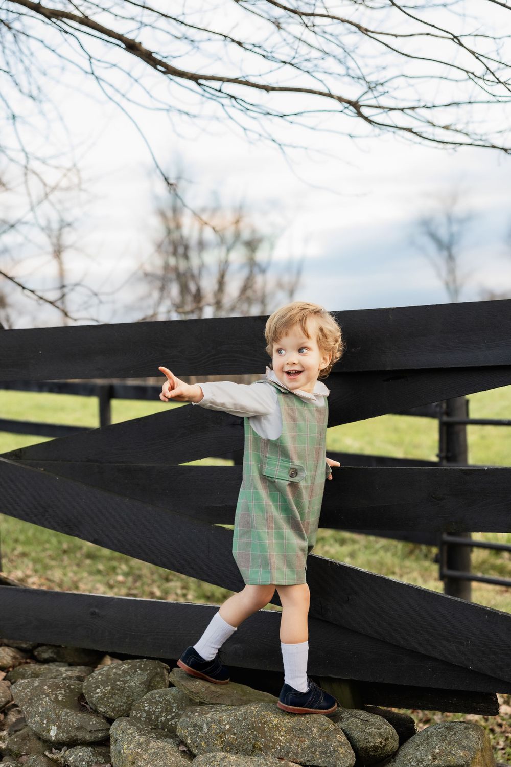 A small child in a plaid dress stands on rocks pointing next to a black wooden fence on a sunny day.