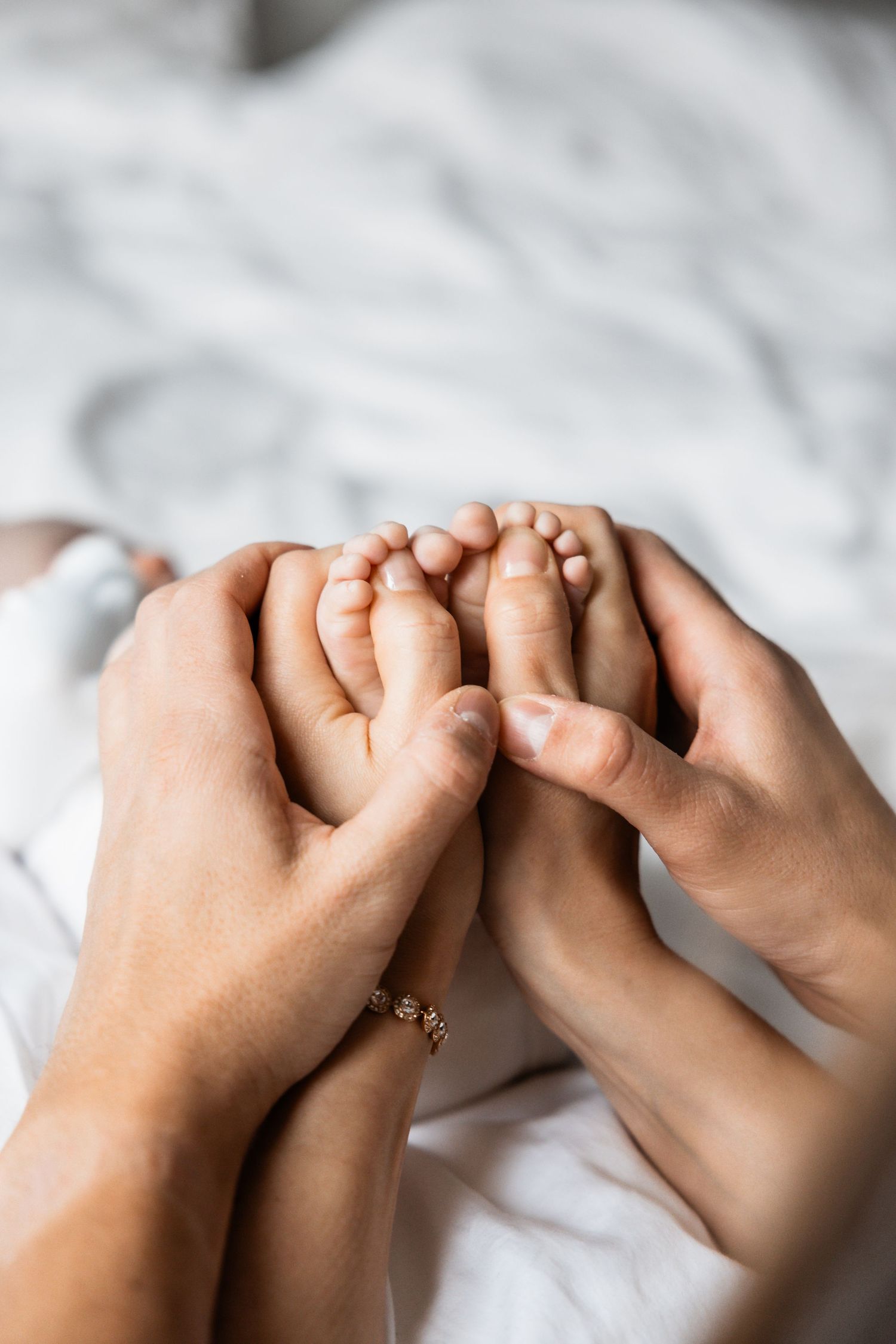 Adult hands gently holding tiny baby feet against white bedding with a delicate bracelet visible.