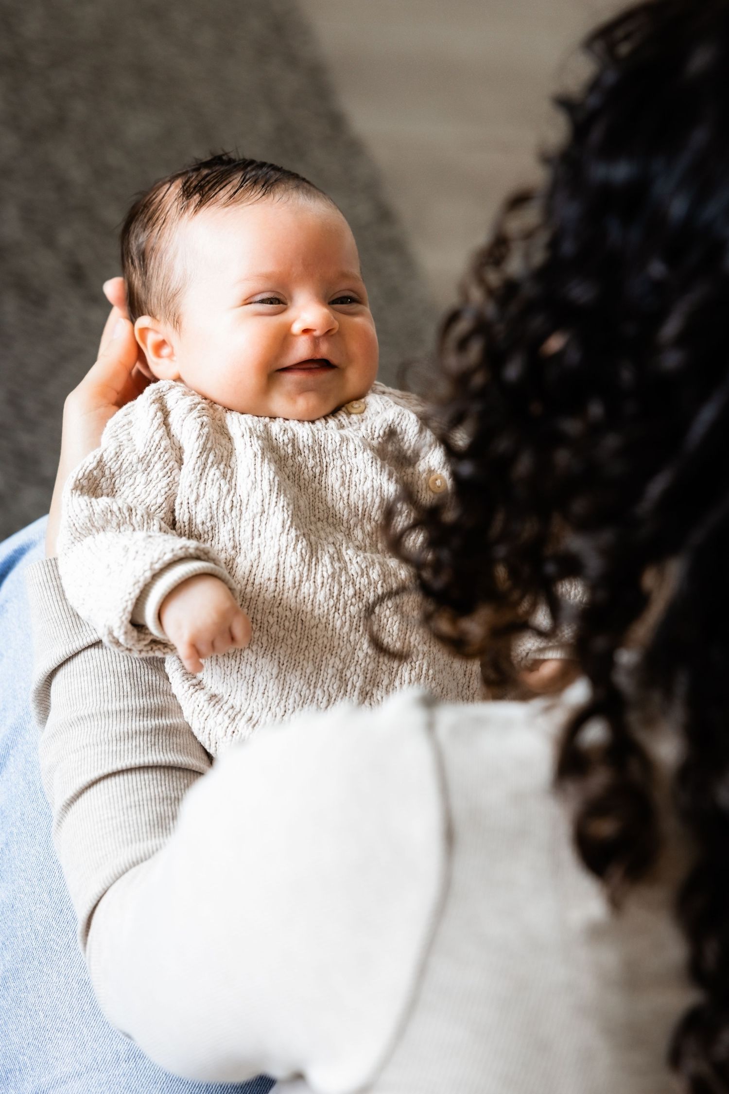 A happy baby wearing a white knit sweater smiles while being held against a light background.