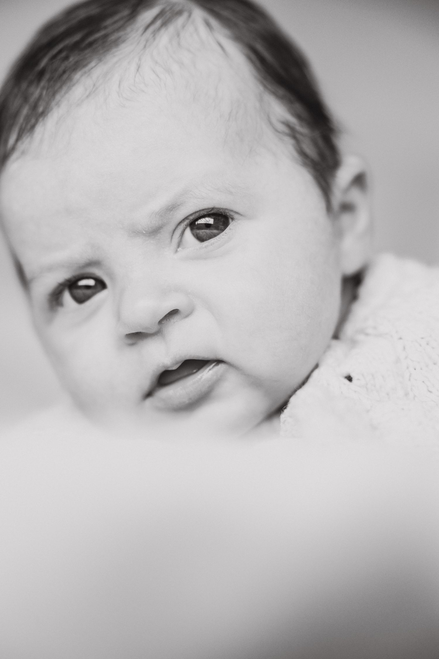 A sweet baby in black and white close-up photograph gazes intently at camera with bright eyes and expressive face.