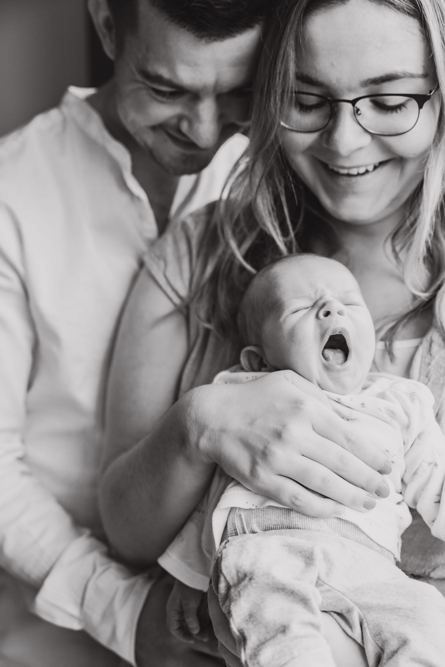 Black and white photo of a couple holding their yawning newborn baby, sharing a tender moment together.