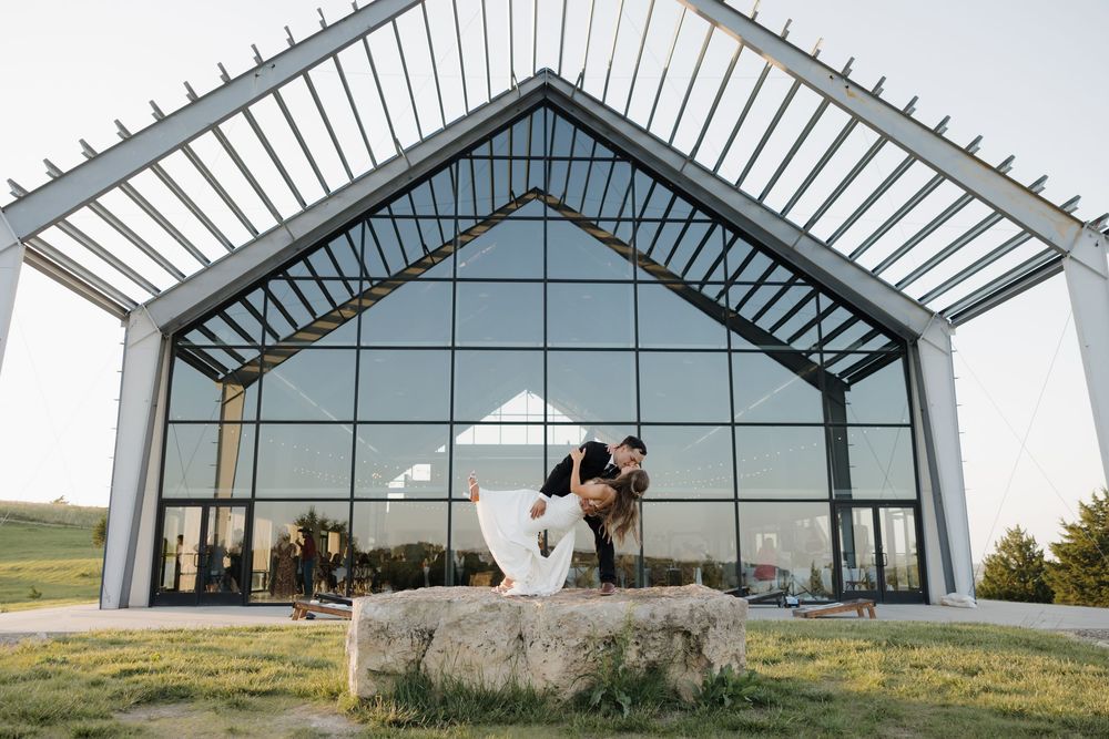 A couple embraces outside a modern chapel with glass walls and angular white architecture at sunset.