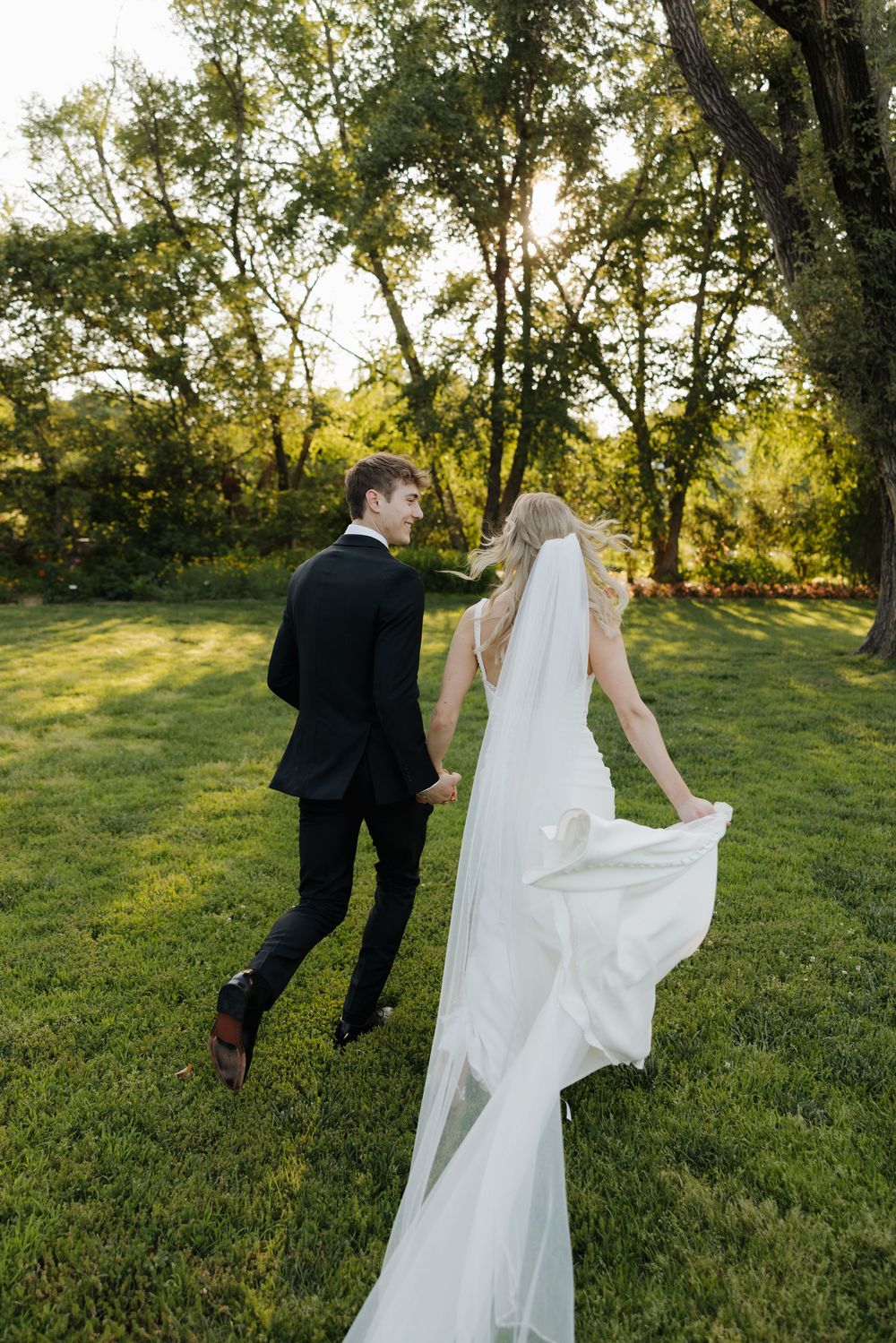 A couple in wedding attire walks hand in hand across a sunlit green field with trees in the background at sunset.