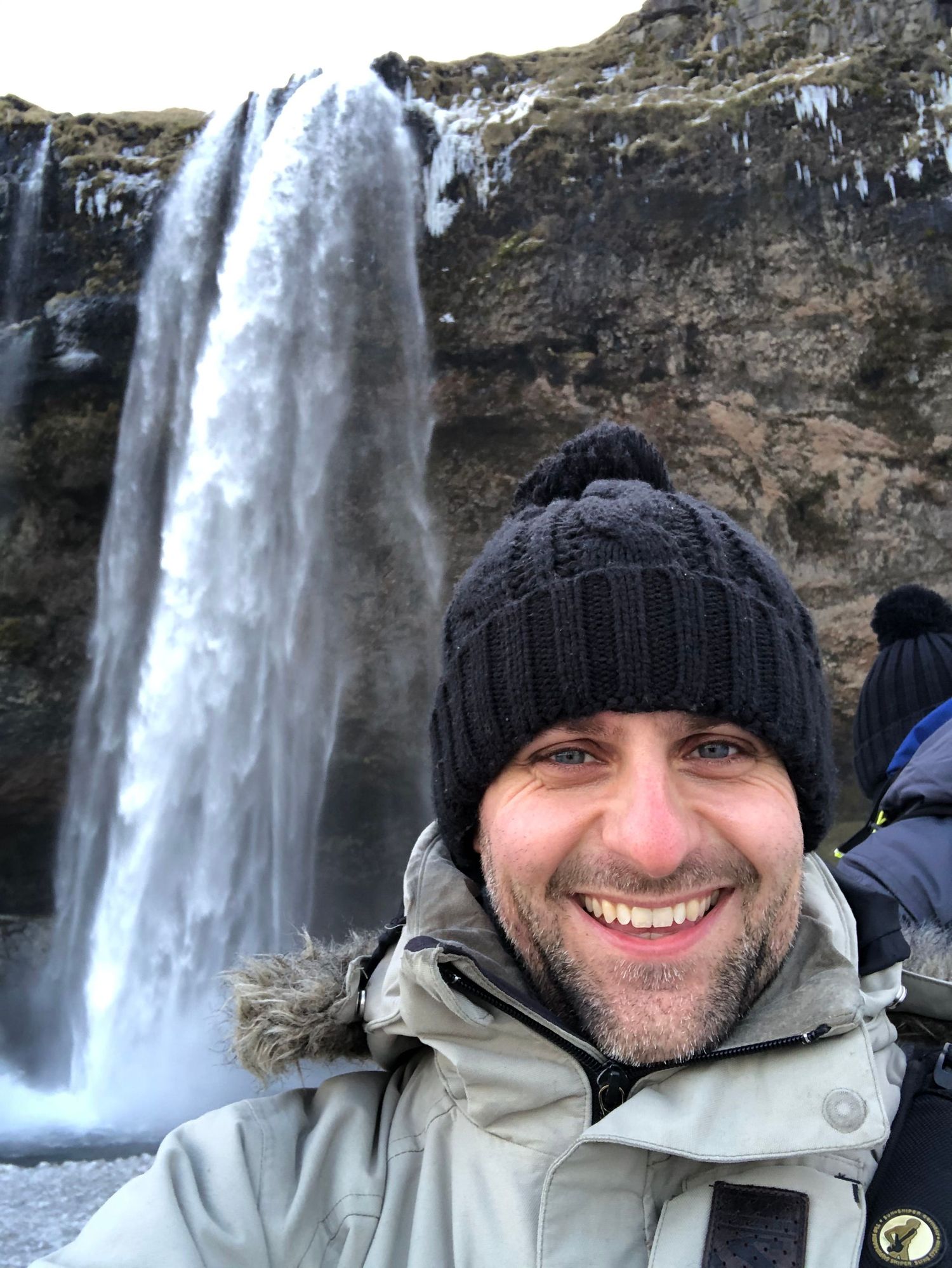 Lee Charlton standing in front of a scenic waterfall cascading down an icy rocky cliff in Iceland with a winter landscape in the background.
