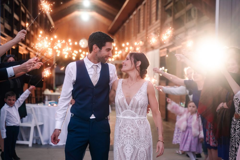 A bride and groom smile at one another as guests hold sparklers behind them at their wedding exit.