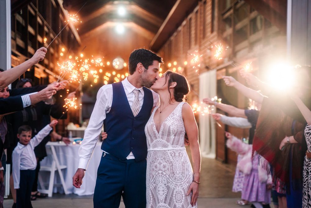 A bride and groom share a kiss as guests hold sparklers behind them.