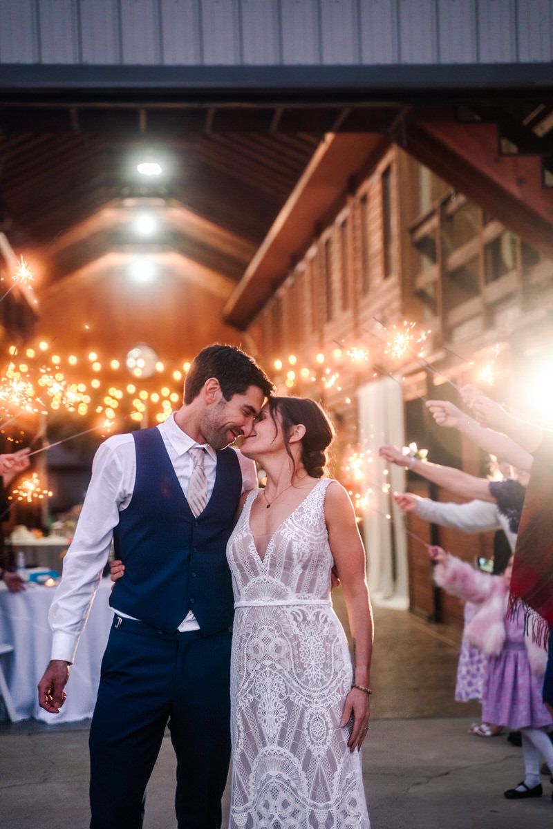 A bride and groom put faces together and smile as guests hold sparklers behind them.