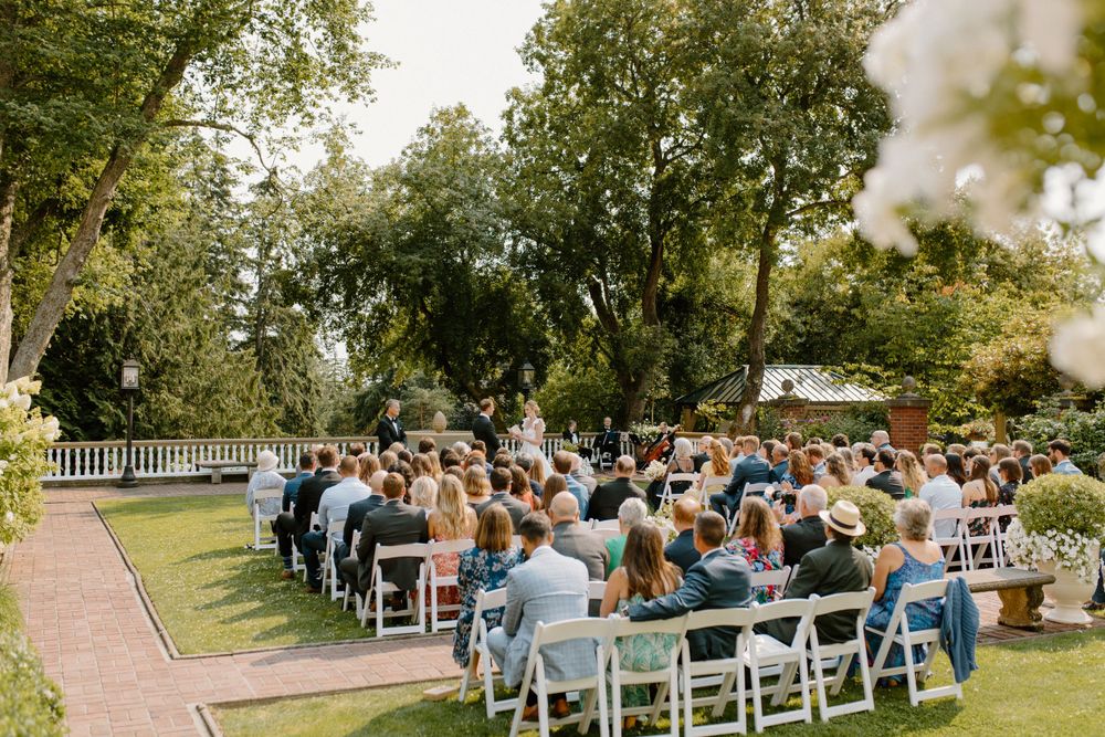 Guests seated in white folding chairs at an outdoor garden wedding ceremony beneath tall green trees.