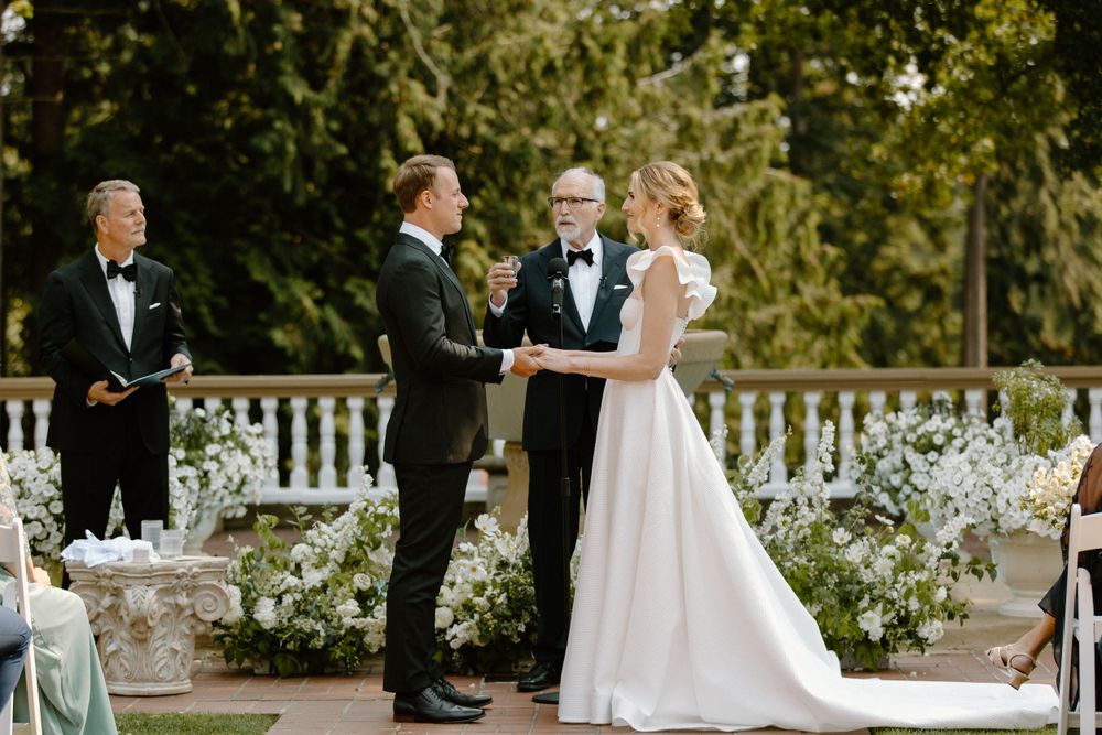 An outdoor wedding ceremony takes place on a scenic terrace surrounded by white floral arrangements and greenery.