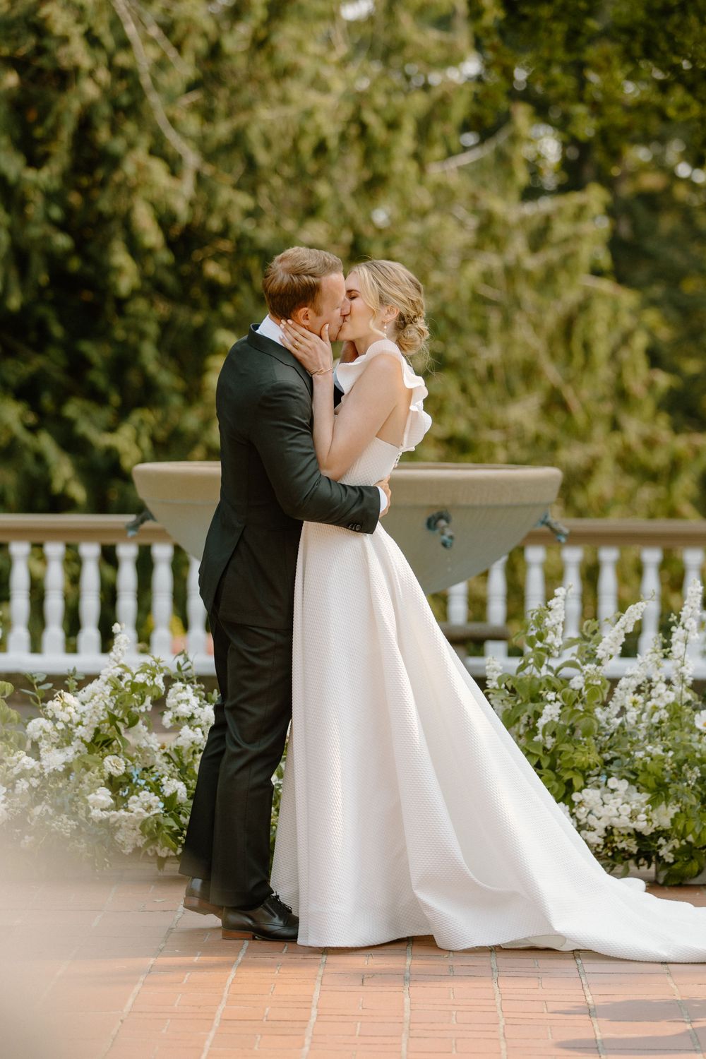Classic wedding moment on a terrace with bride in elegant white gown and groom in black suit embracing against greenery backdrop.
