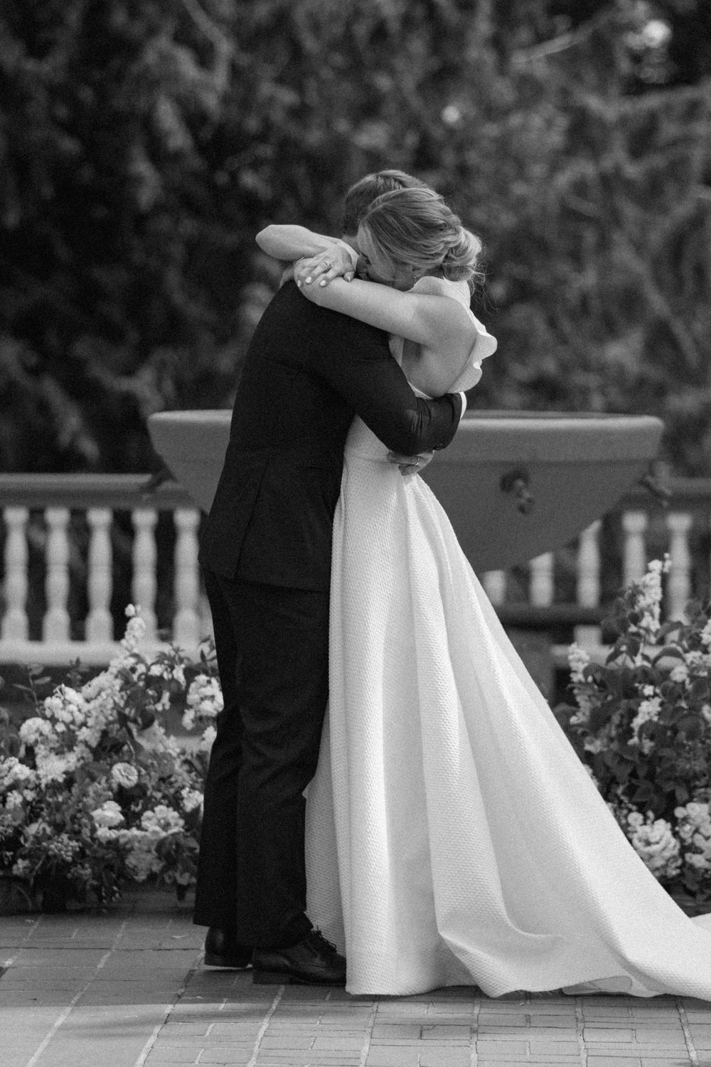 A black and white wedding photo of a couple embracing on a terrace surrounded by flowers and decorative railings.