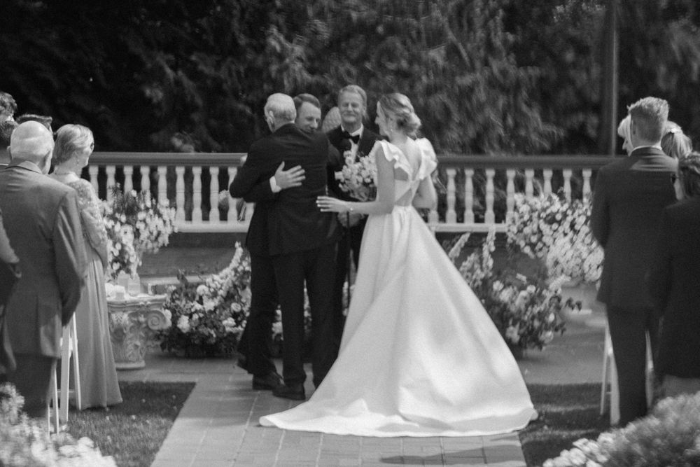 Black and white photograph of a wedding ceremony taking place outdoors with guests gathered around a decorated garden setting.