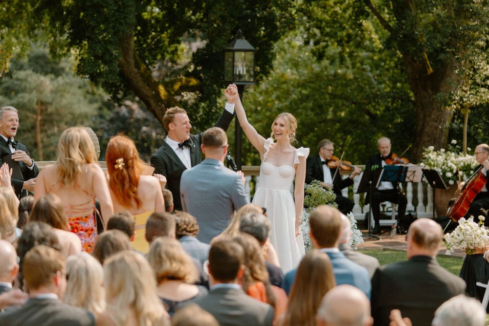 Outdoor garden wedding ceremony with guests seated in rows under trees during golden sunlight hours.