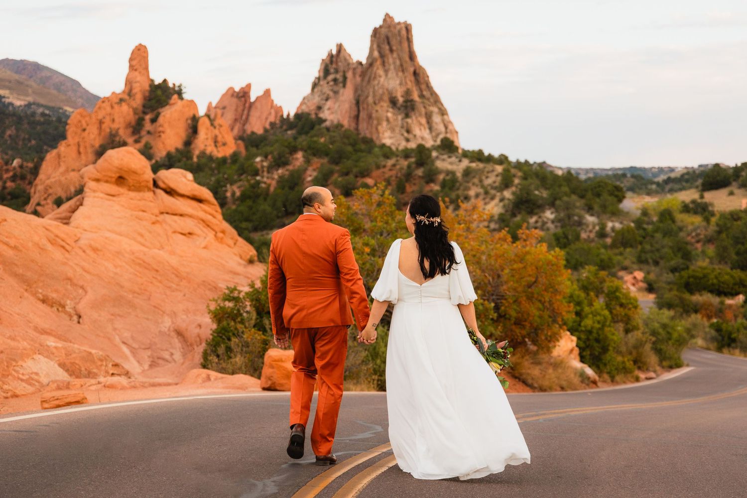 A couple in wedding attire walk along a scenic road at Garden of the Gods park with red rock formations at sunset.