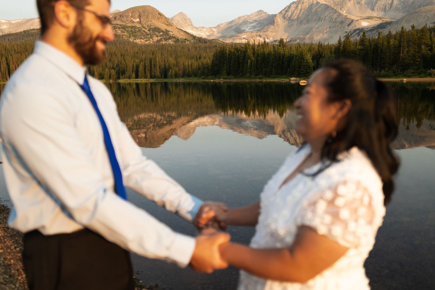 A couple holds hands during their outdoor mountain wedding ceremony beside a serene lake at sunset.