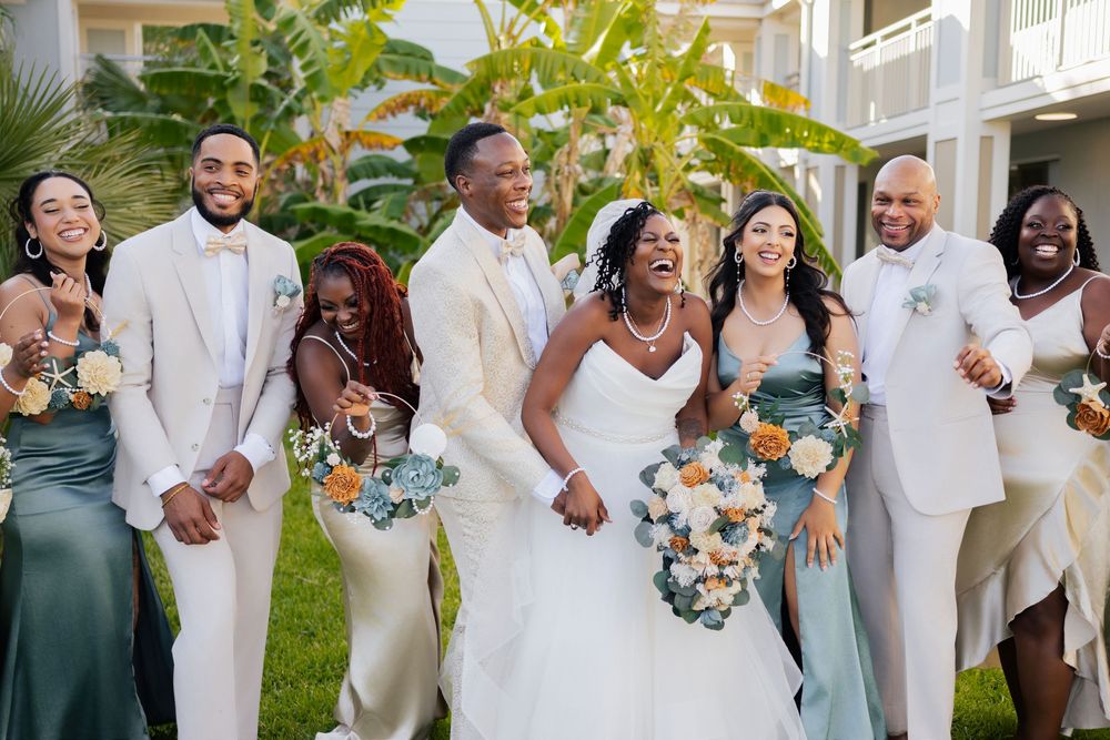 A wedding party in elegant white and sage green attire poses together on a lawn with tropical palm trees in the background.