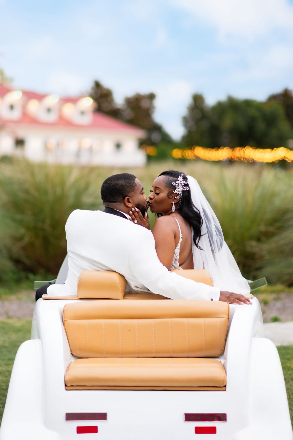 A newlywed couple shares a romantic moment while riding in a white golf cart with tan leather seats.