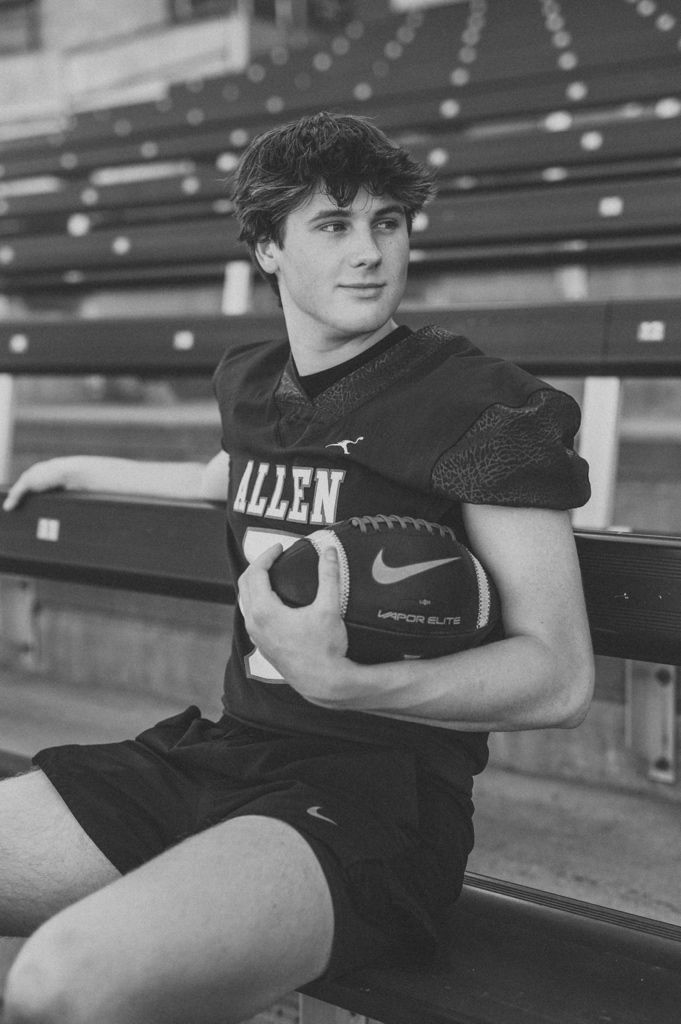 High school football athlete in black Allen jersey holds football while sitting on stadium bleachers in black and white photo.