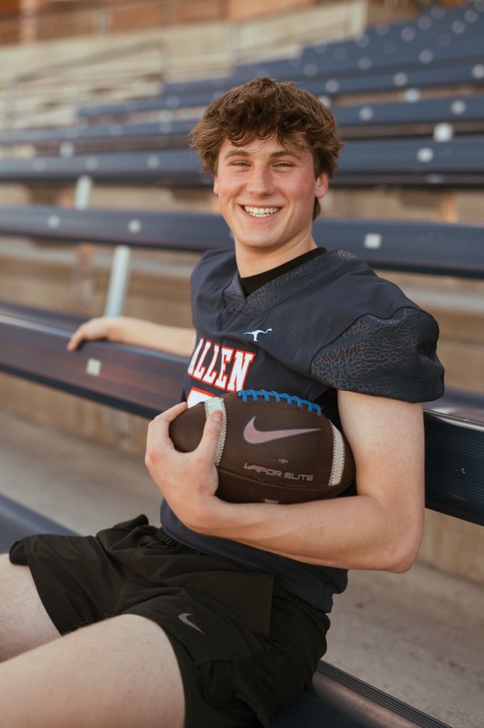 High school football player in uniform holds football while sitting on empty stadium bleachers during photo session.