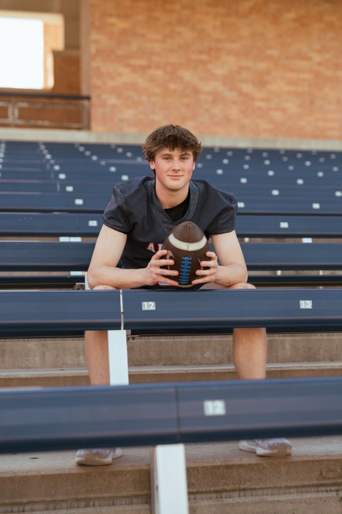 Person in black shirt sits on stadium bleachers looking forward while holding a football in their hands.