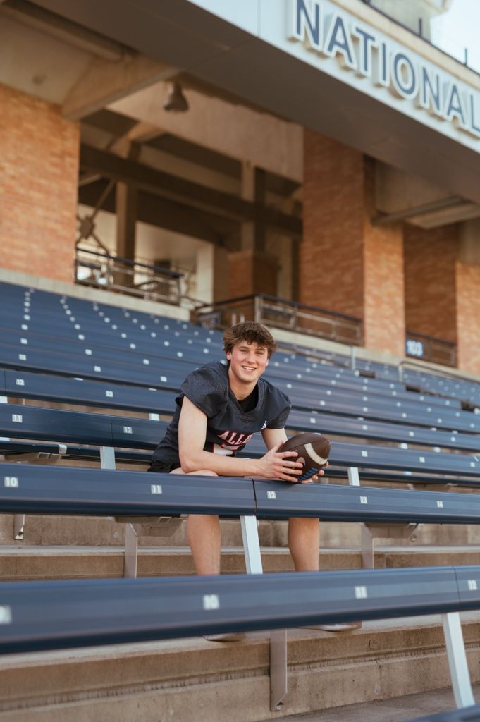A student athlete sits on stadium bleachers wearing a black t-shirt while holding a football at National Ballpark.