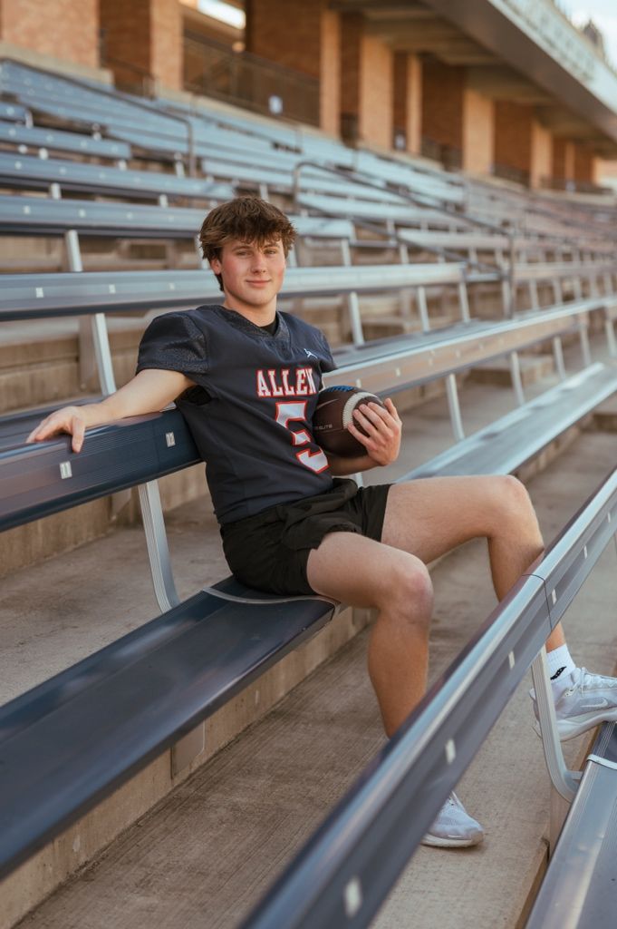 Football player in a black jersey and shorts sits on bleachers holding a ball at a stadium during golden hour.