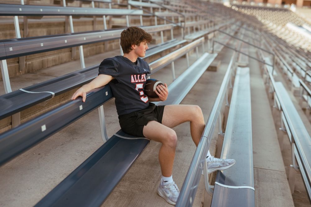 Young athlete in black Illinois t-shirt sits alone on empty bleacher seats at sports stadium holding basketball.