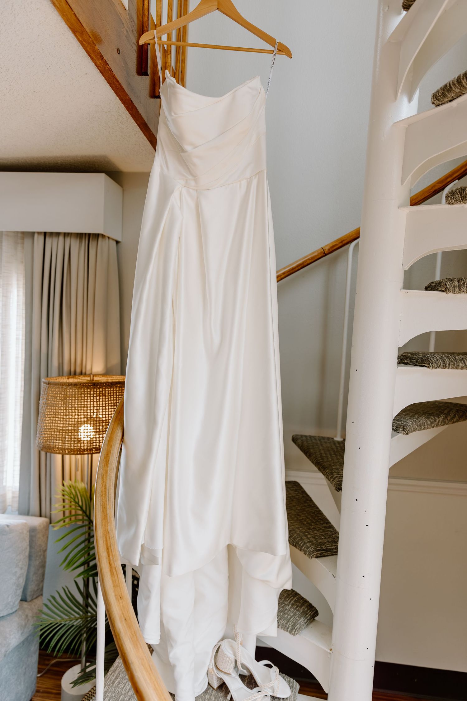 A white wedding dress hangs on a wooden hanger against a spiral staircase wall with a rattan lamp in the background.