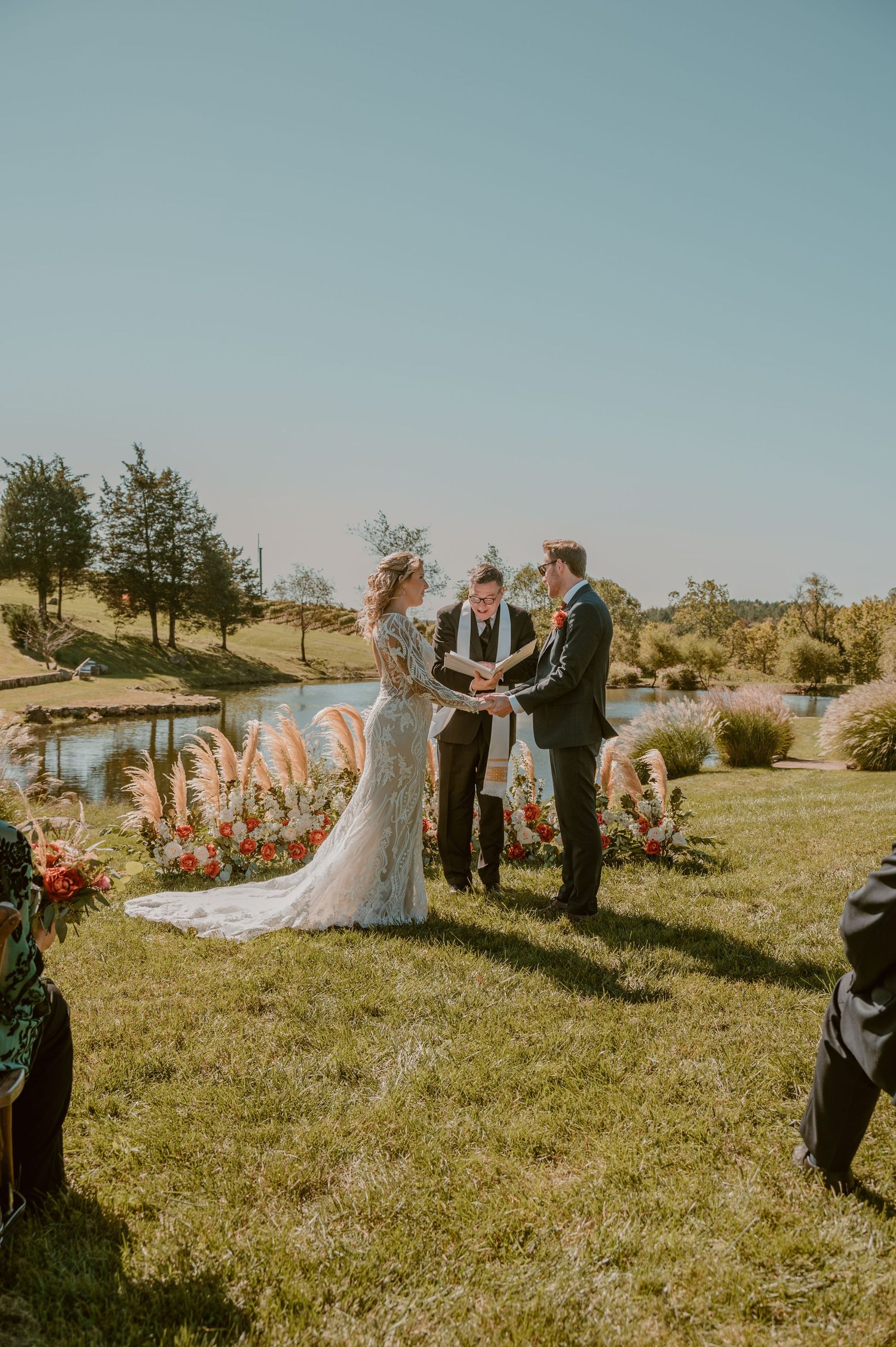 An outdoor wedding ceremony takes place on a grassy hill overlooking a pond on a sunny day with floral decorations. by Samantha Campbell