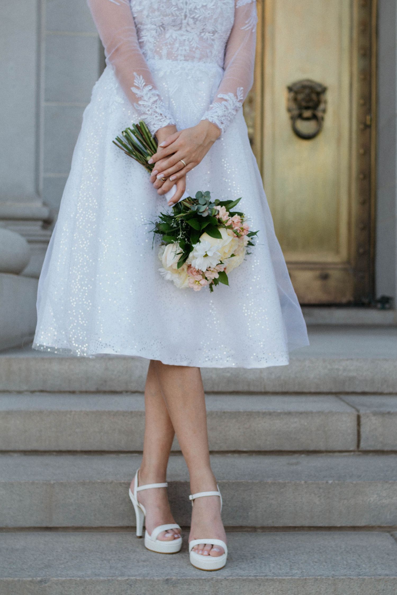 Bride in tea-length lace wedding dress and white sandals holds bouquet of white flowers on stone steps. Elopement, Denver Colorado