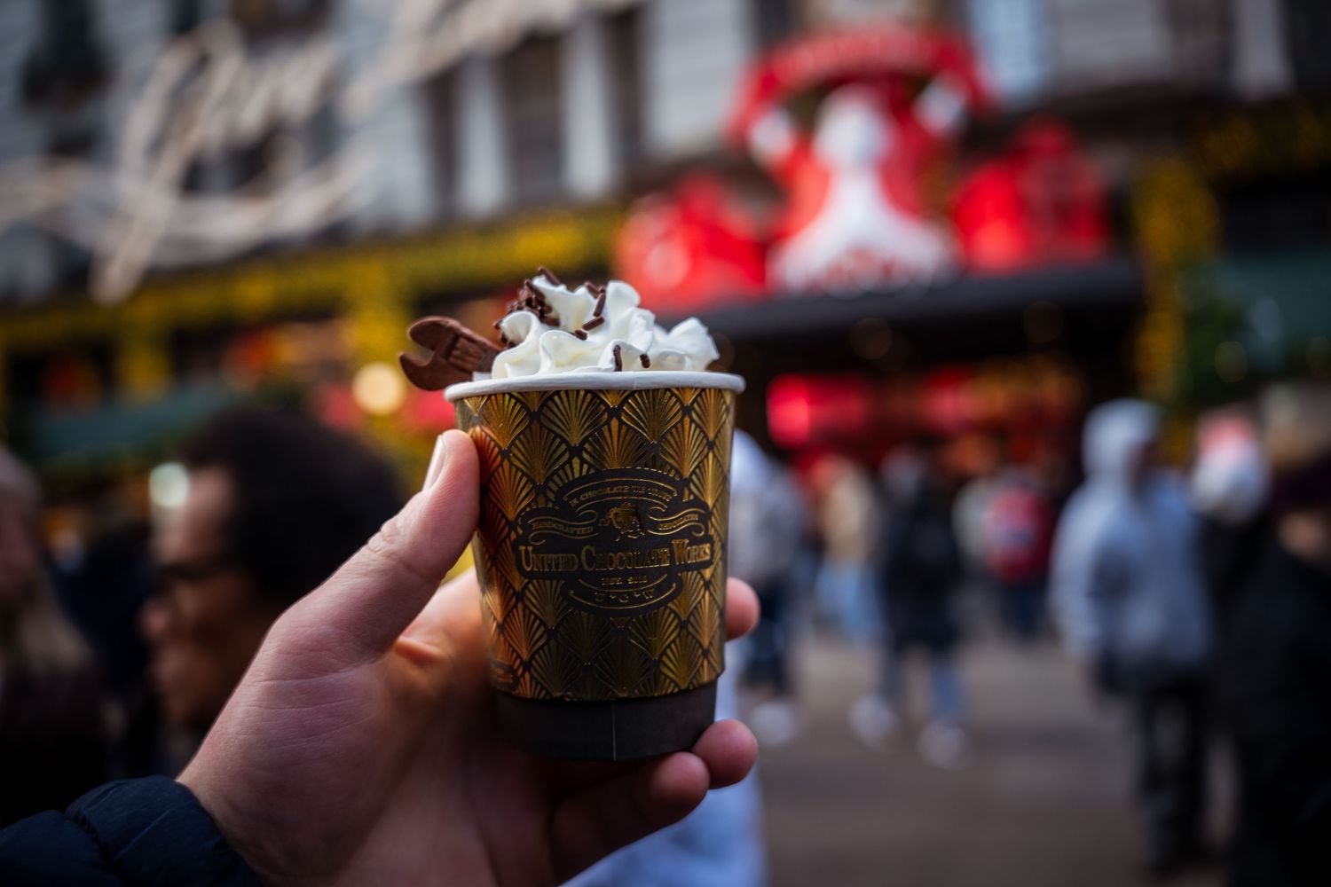 Hand holding a hot chocolate, with a festive Christmas display softly blurred in the background.