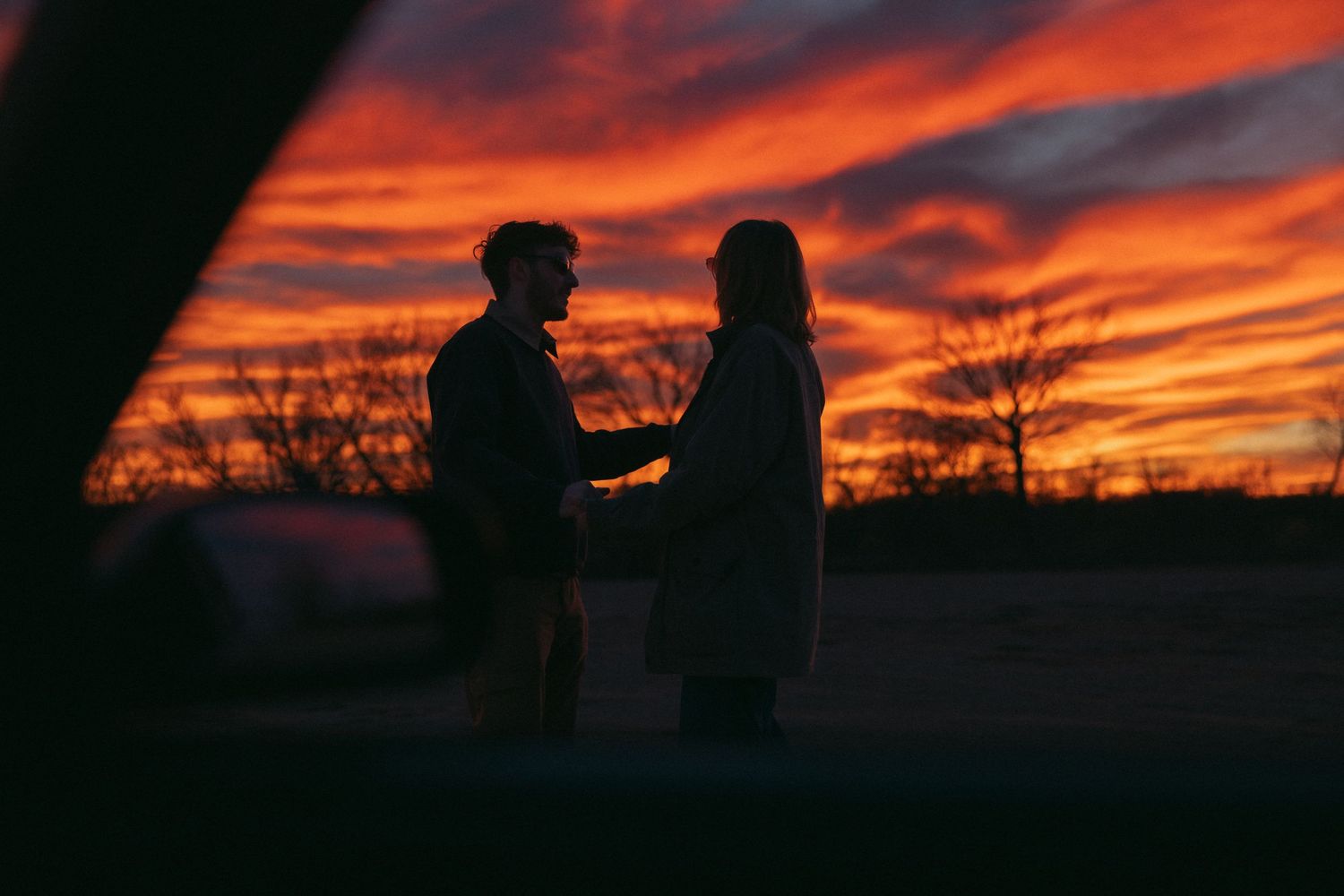 Two silhouettes stand together against a vibrant orange and red sunset sky with bare tree branches in the background.