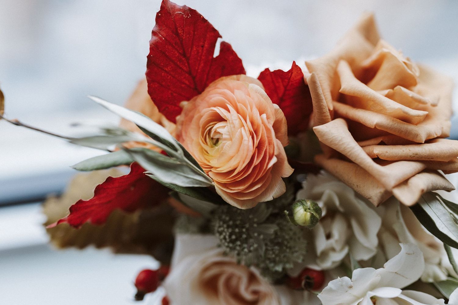 Elegant wedding bouquet featuring blush ranunculus, peach roses and deep red autumn leaves in soft focus.