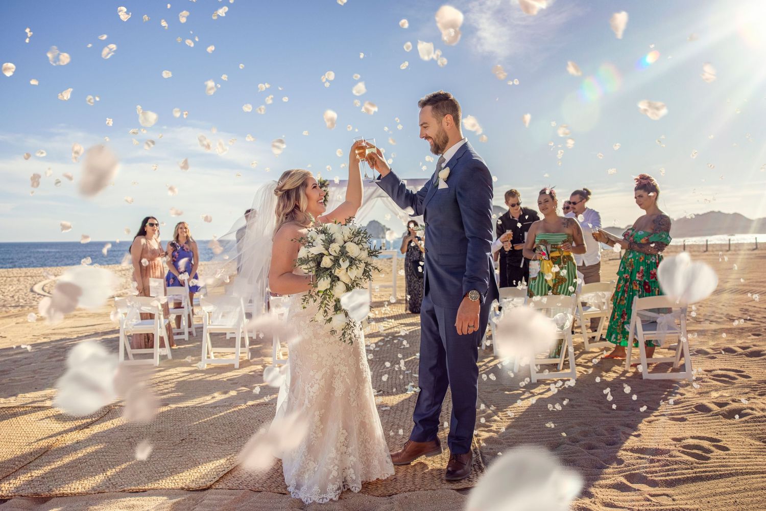 A beach wedding ceremony with flower petals floating through the air as newlyweds celebrate on a sunny day by the ocean.