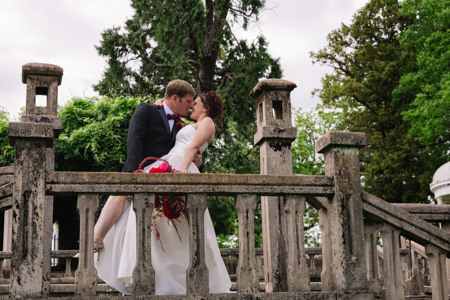 A romantic couple in wedding attire share an intimate moment on a rustic stone bridge surrounded by lush greenery. Bleak House, Tennessee.